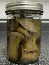 Load image into Gallery viewer, A clear glass mason jar with a silver metal lid, filled with sliced mushrooms submerged in a liquid, photographed on a dark countertop.
