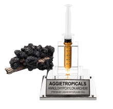 تحميل الصورة في عارض المعرض، A clear acrylic and glass syringe-style container filled with amber liquid, mounted on a silver base engraved with 'AGGJETROPICALS ANNULOHYPXYLON ARCHERI PREMIUM LIQUID MYCELIUM 10cc'. A black fungal cluster sits to the left on a white background.
