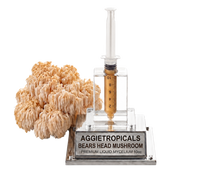 A bottle-shaped specimen display for Aggietropicals Bears Head Mushroom, featuring a clear plastic syringe filled with golden-brown liquid mounted on a clear acrylic stand with a silver base plate, next to a beige honeycomb-like mushroom cluster on a white background.