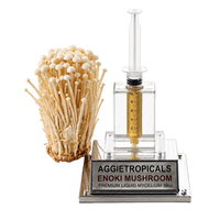 A clear acrylic enoki mushroom mycelium kit by Aggietropicals, featuring a liquid culture chamber with a syringe-style measurement device and a clear glass bottle containing golden liquid, shown next to a cluster of mature beige enoki mushrooms on a silver base.