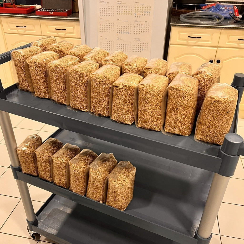 A three-tier gray utility cart loaded with multiple clear plastic bags filled with a tan-brown granular substance, photographed in a kitchen environment.