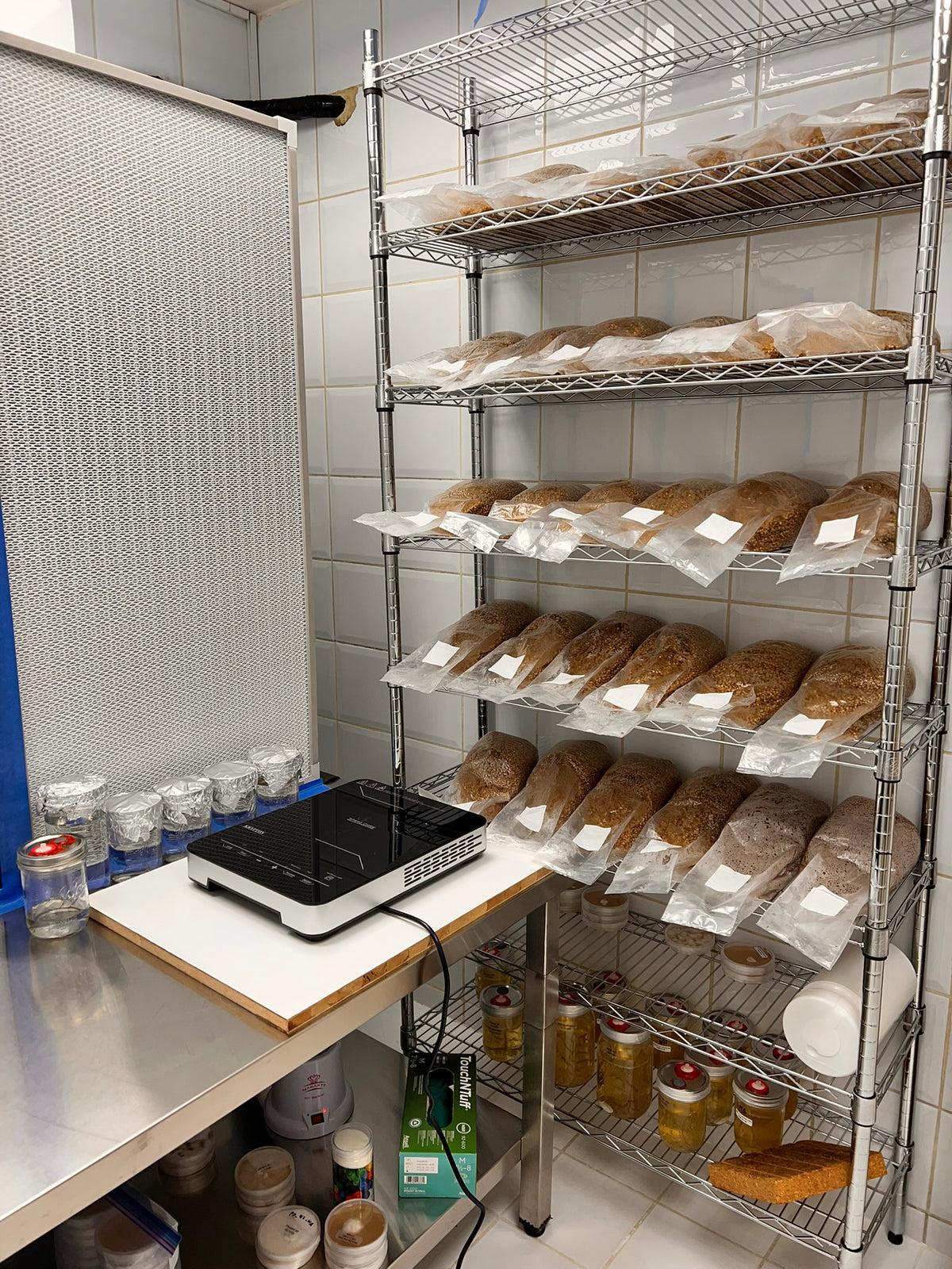 A stainless steel countertop with a black and white digital scale on a white cutting board, next to a large white mesh panel. To the right are tall wire shelving units stocked with numerous clear plastic bags of brown and beige dried or ground ingredients, with glass jars and bottles on the lower shelf.