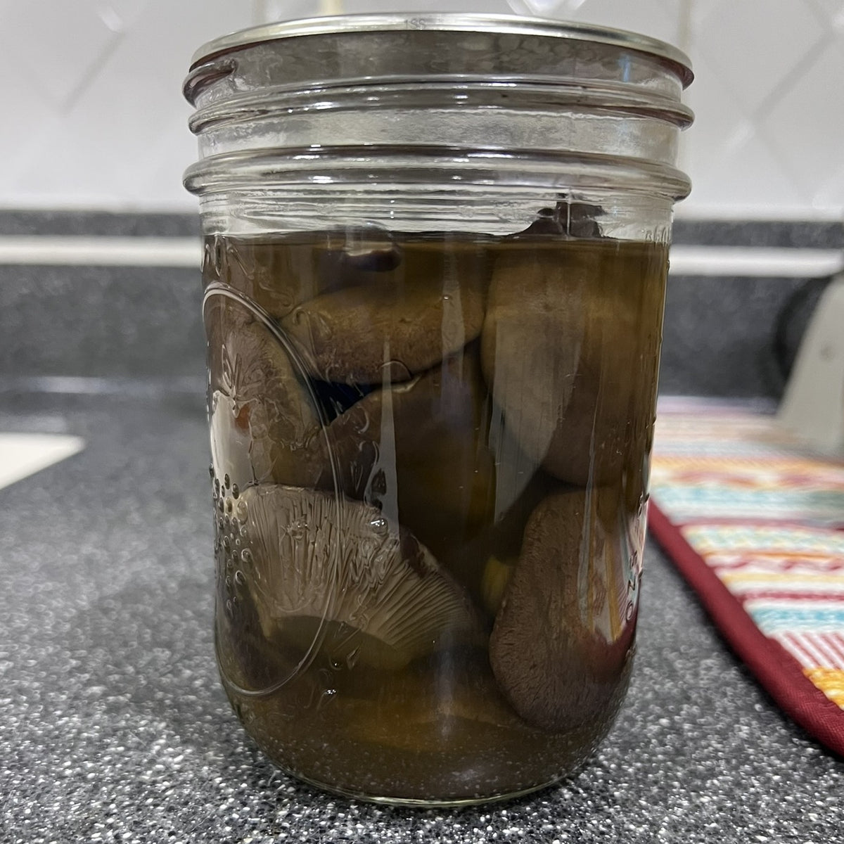 A clear glass mason jar with a metal lid, filled with sliced mushrooms submerged in a dark liquid, sitting on a speckled gray countertop in a kitchen setting.