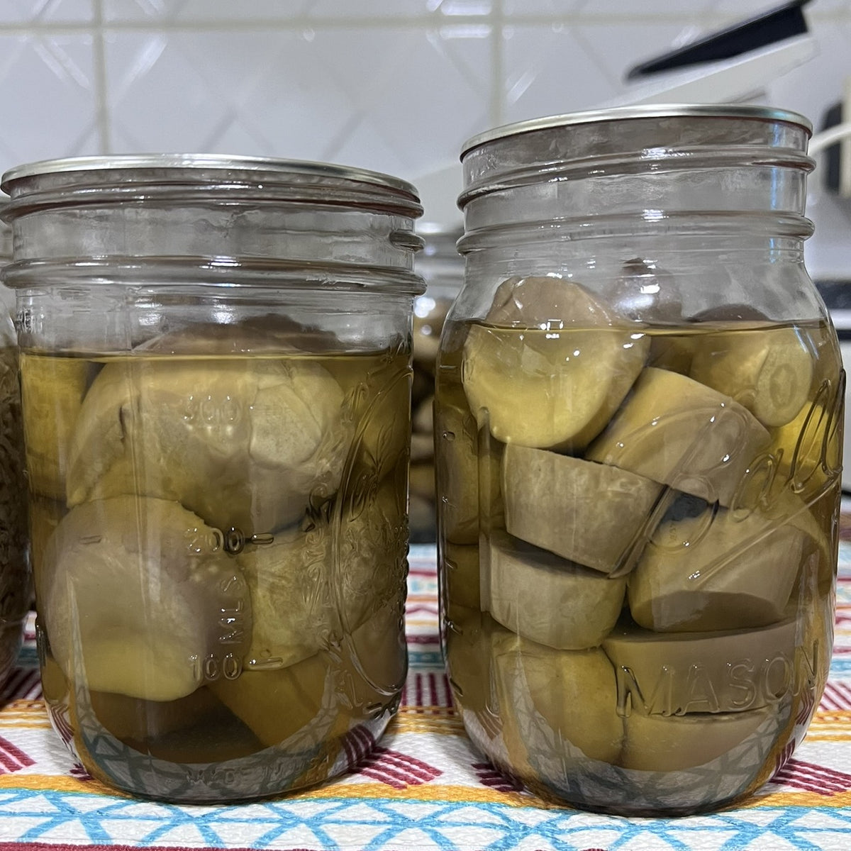 Two glass mason jars filled with sliced mushrooms soaking in liquid, set on a patterned tablecloth with a tiled wall in the background.