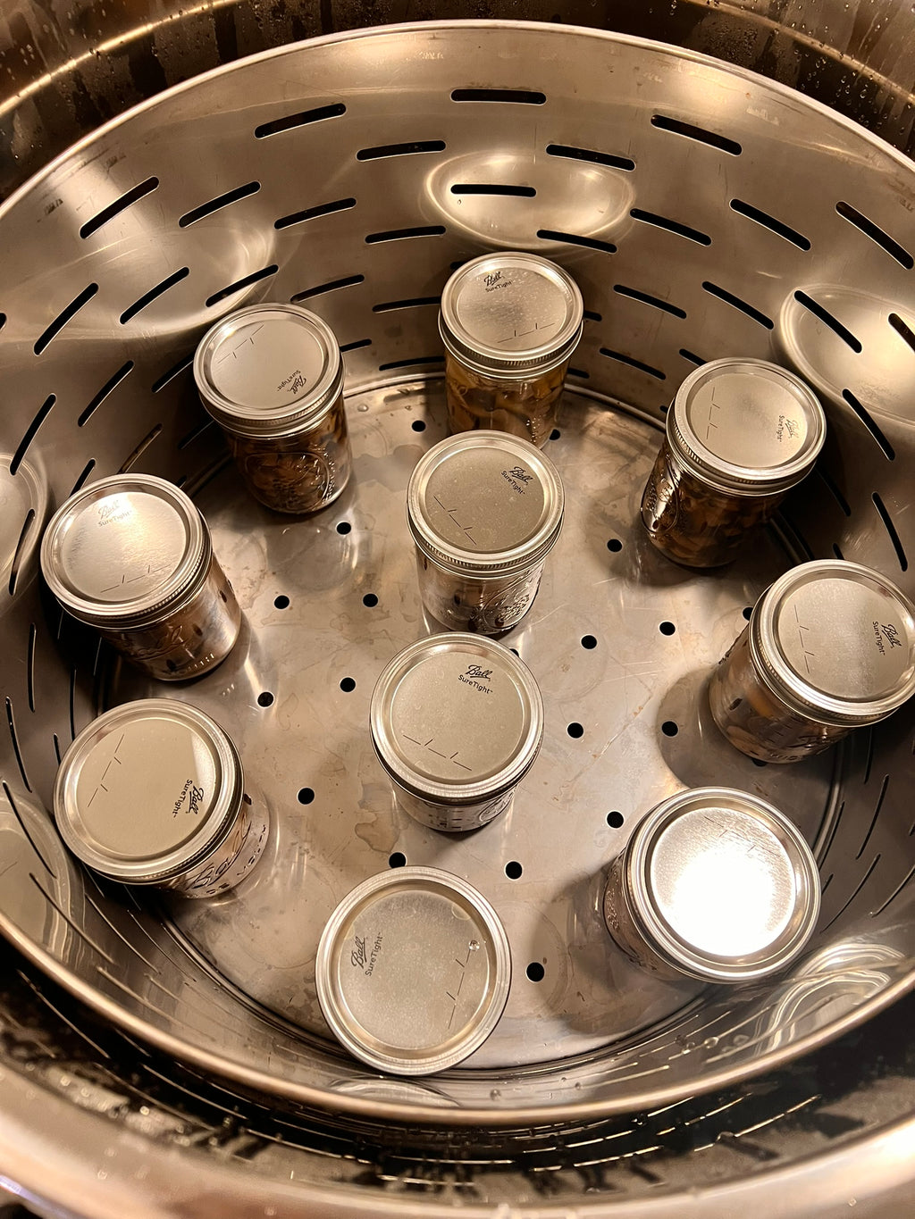 Several small glass jars with silver metal lids arranged inside a stainless steel canner steamer basket.