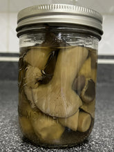 Load image into Gallery viewer, A clear glass mason jar with a silver metal lid, filled with sliced mushrooms submerged in liquid, photographed on a dark speckled countertop with a tiled wall in the background.
