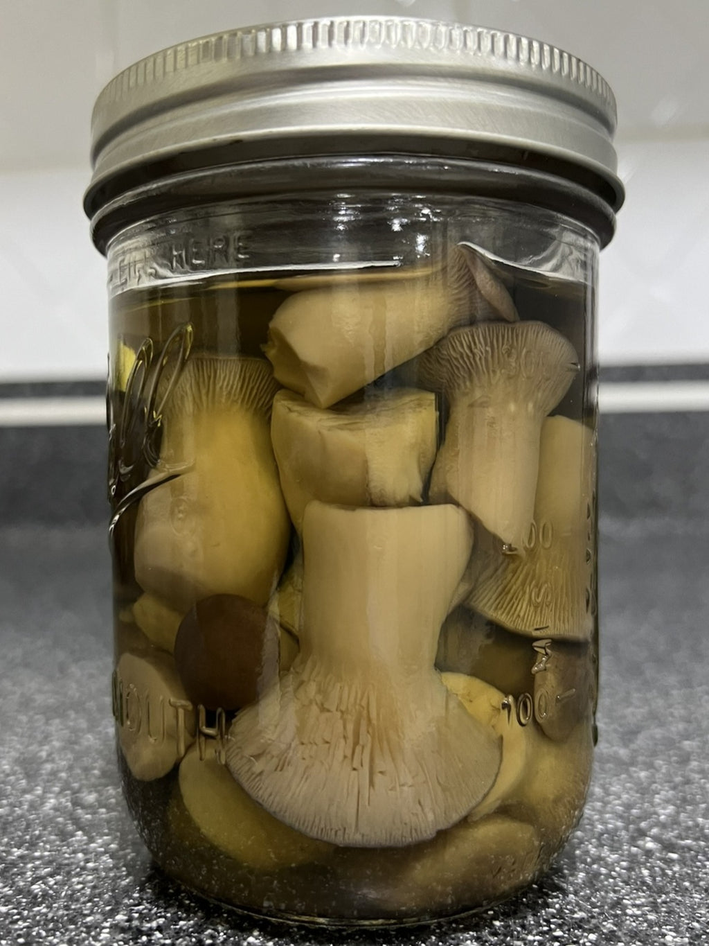 A clear glass mason jar with a silver metal lid, filled with sliced mushrooms submerged in a liquid, photographed on a dark countertop.