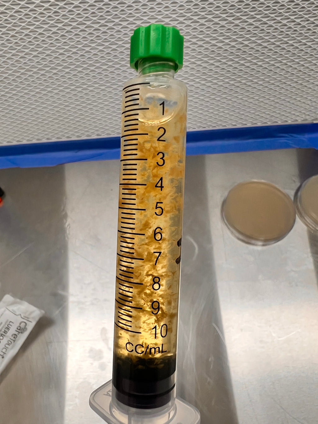 A clear plastic syringe-style container with measurement markings in mL, filled with a yellowish liquid and containing small floating mycelium particles, with a green cap, photographed on a metallic surface.