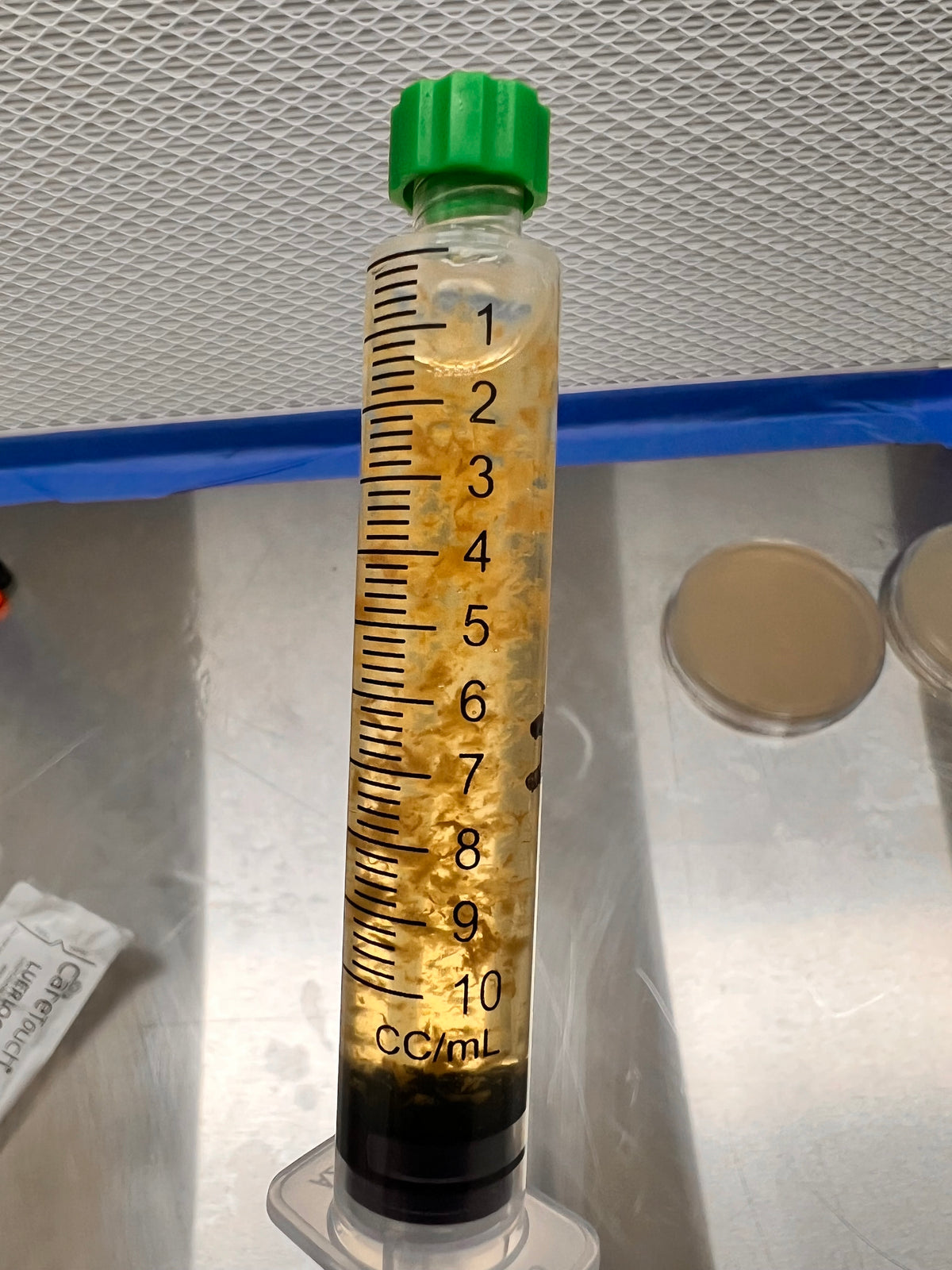 A clear plastic syringe-style container with measurement markings in mL, filled with a yellowish liquid and containing small floating mycelium particles, with a green cap, photographed on a metallic surface.