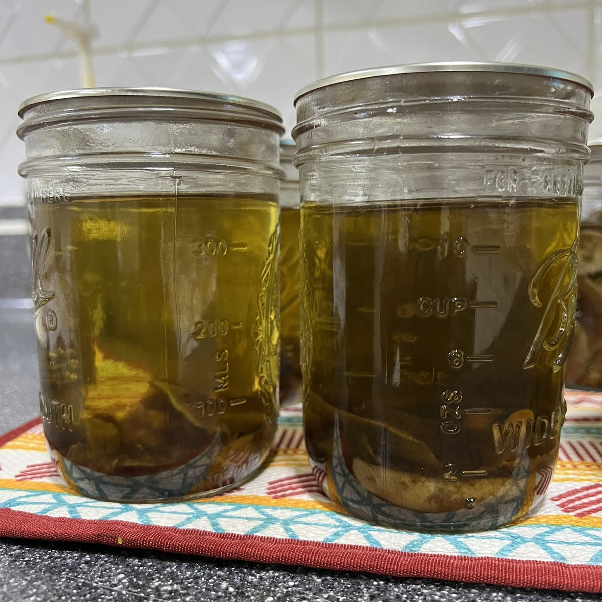 Two glass mason jars filled with an amber-colored liquid, likely infused mushroom or tincture, sitting on a patterned kitchen towel on a countertop.
