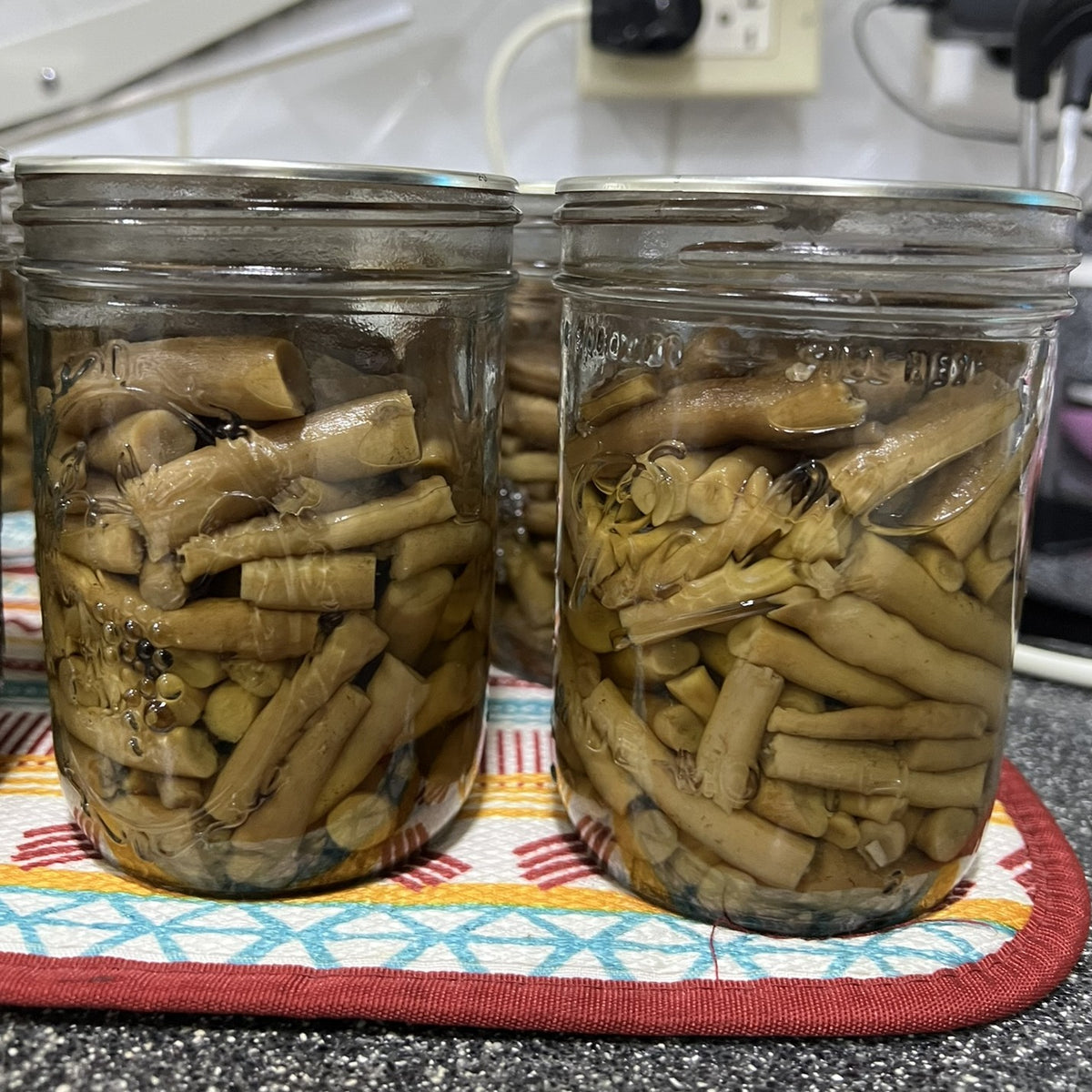 Two glass mason jars filled with sliced pioppino mushroom stems in liquid, placed on a patterned trivet on a kitchen countertop.