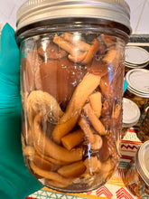 Load image into Gallery viewer, A clear glass mason jar filled with sliced mushrooms submerged in liquid, held by a teal gloved hand. More jars are visible in the background on a patterned surface.
