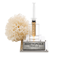 A 10ml syringe filled with golden liquid mycelium displayed in a clear acrylic holder on a silver metal base engraved with 'AGGIETROPICALS TREMELLA PREMIUM LIQUID MYCELUM 10cc'. A natural white sponge is positioned to the left of the syringe on a clean white background.